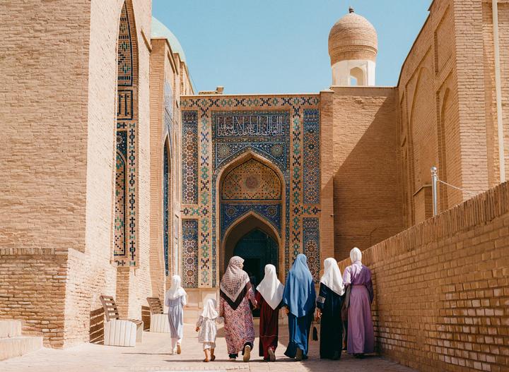 A group of women and children walk towards a building with intricate blue and gold tile work under a clear sky.
