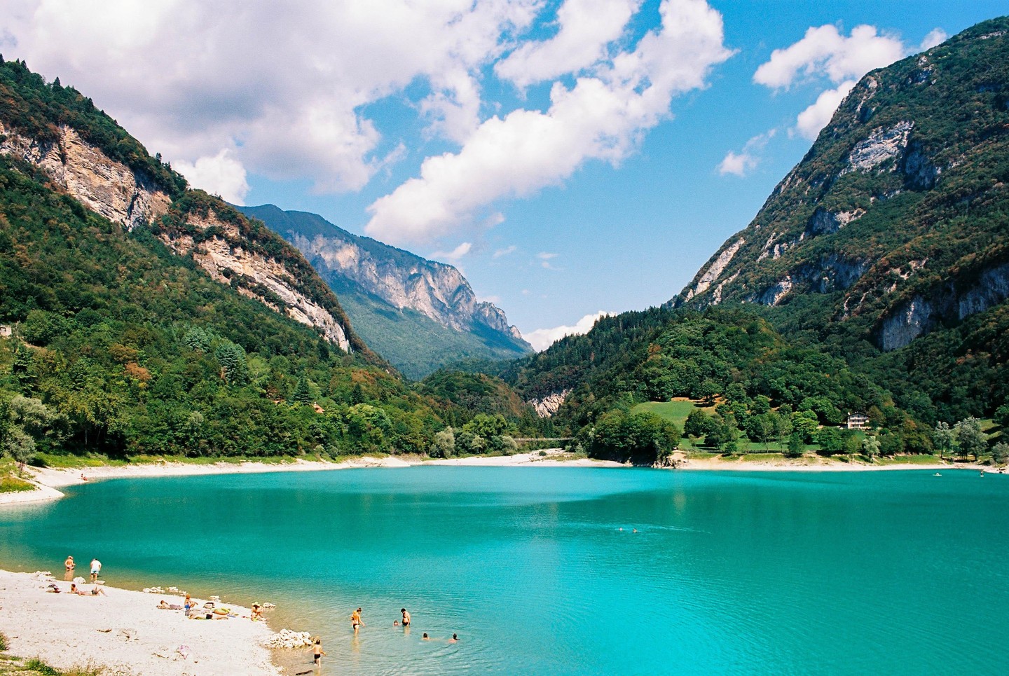 A turquoise lake is surrounded by mountains and a small beach with people swimming and sunbathing.