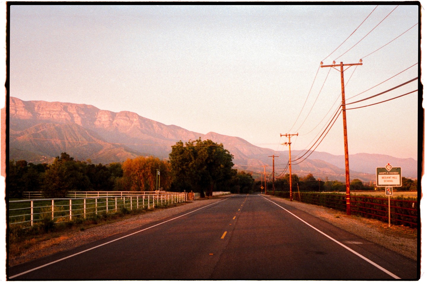A road leads towards mountains at dusk, with power lines and a white fence on either side.