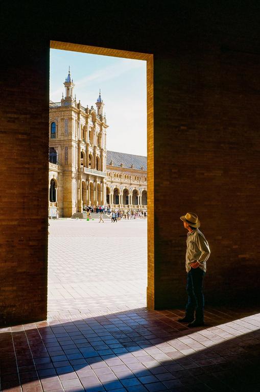 A man in a hat stands in shadow looking out through a doorway at a large ornate building and people in a sunny plaza.