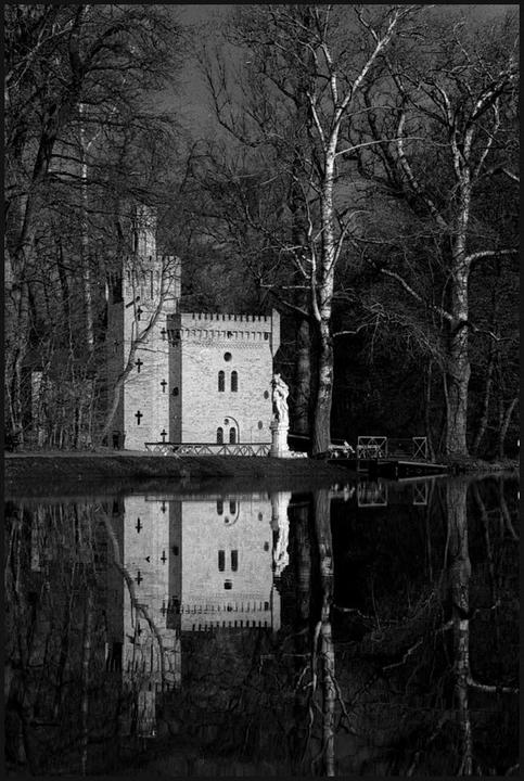 A small castle with a tower and a statue stands by a body of water, surrounded by bare trees, with its reflection visible in the water.