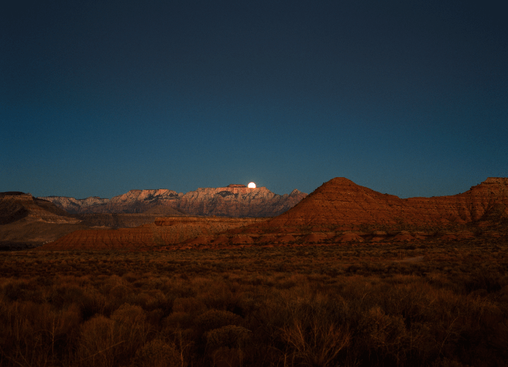 A full moon rises over a desert landscape with mountains and scrub brush under a dark blue sky.
