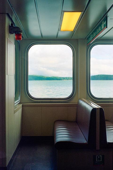 A ferry interior with a brown bench seat and windows looking out onto a body of water with a forested shore.