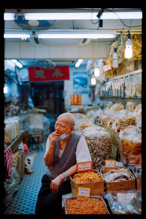 A man with a face mask under his chin smokes a cigarette while sitting in a market stall surrounded by dried goods.