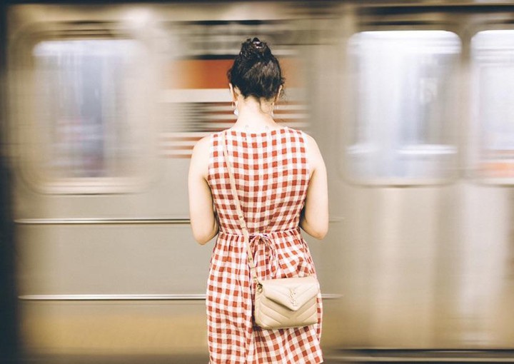 A woman in a red and white checkered dress stands on a subway platform as a train passes by.