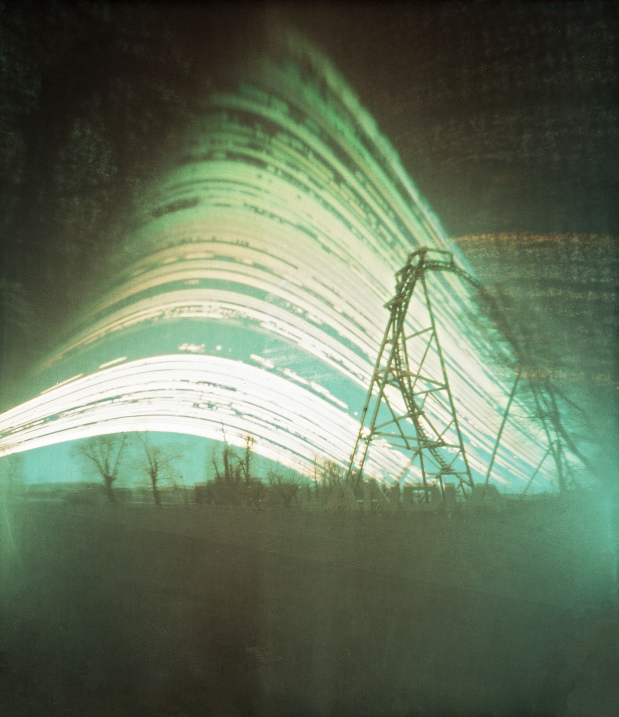 A long exposure photograph captures light trails arcing over a power line tower and a distant treeline.