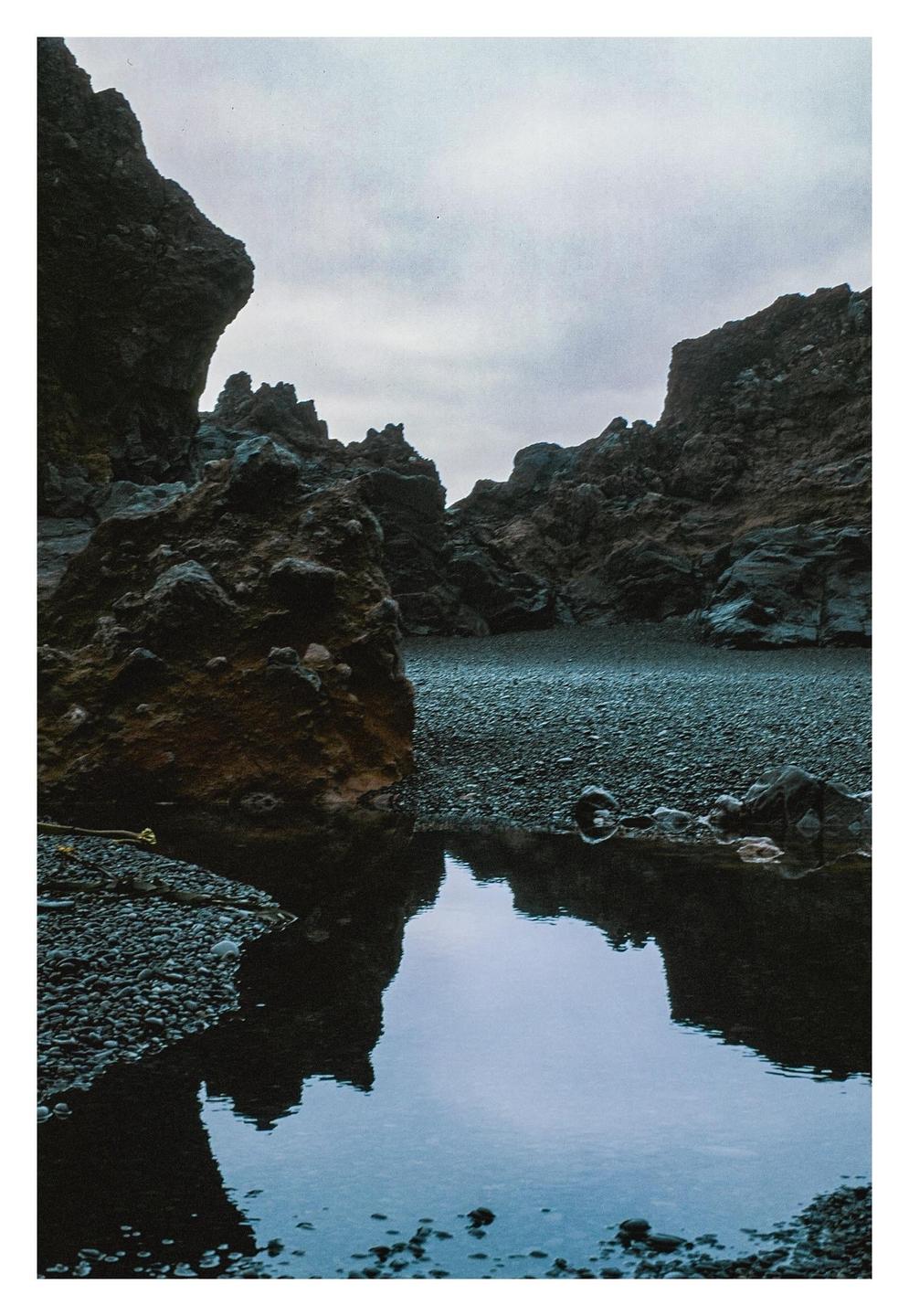 A pool of water reflects the sky and surrounding rock formations on a rocky beach.