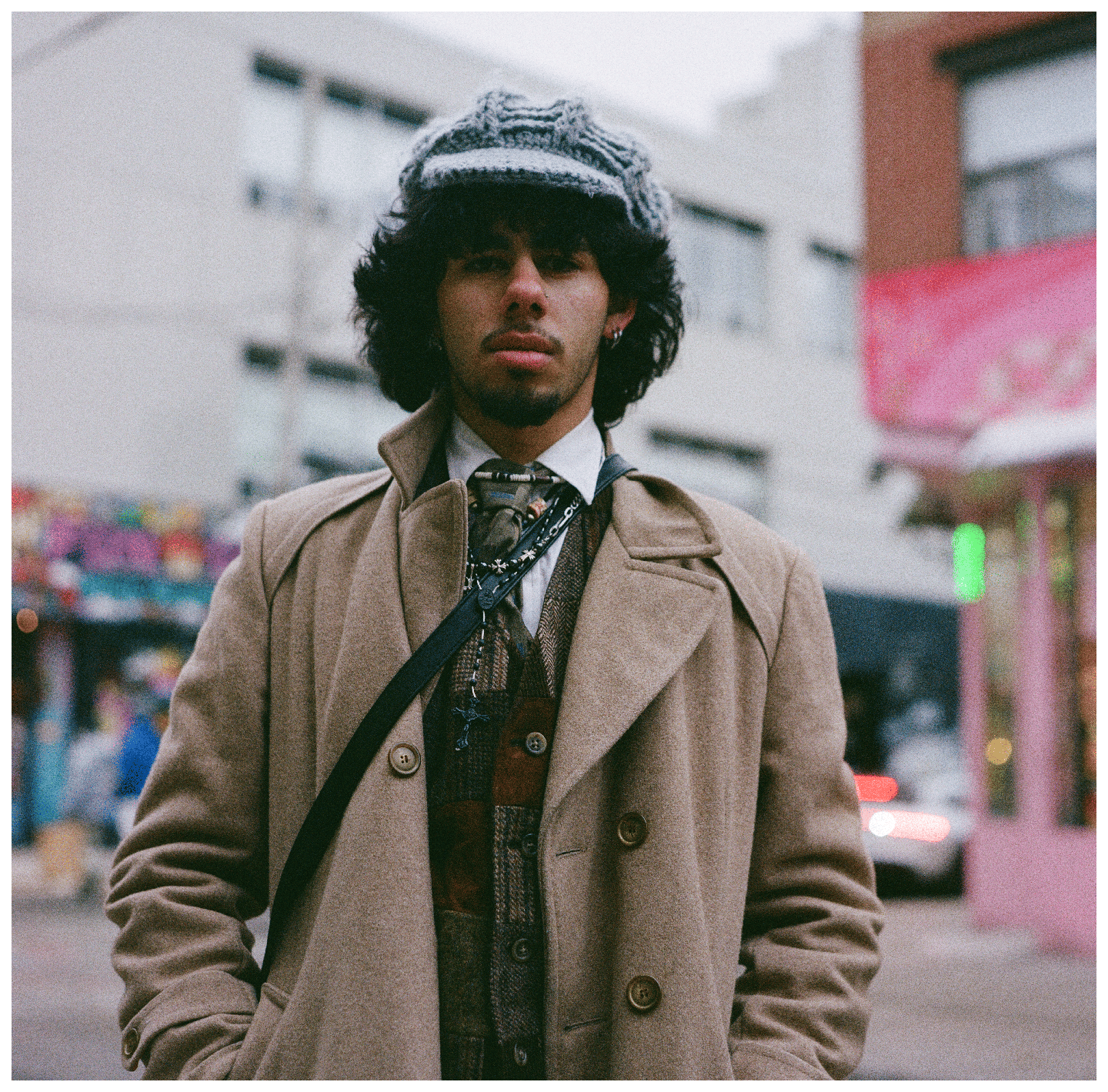 A man wearing a trench coat, a patterned vest, and a knitted cap stands on a street.