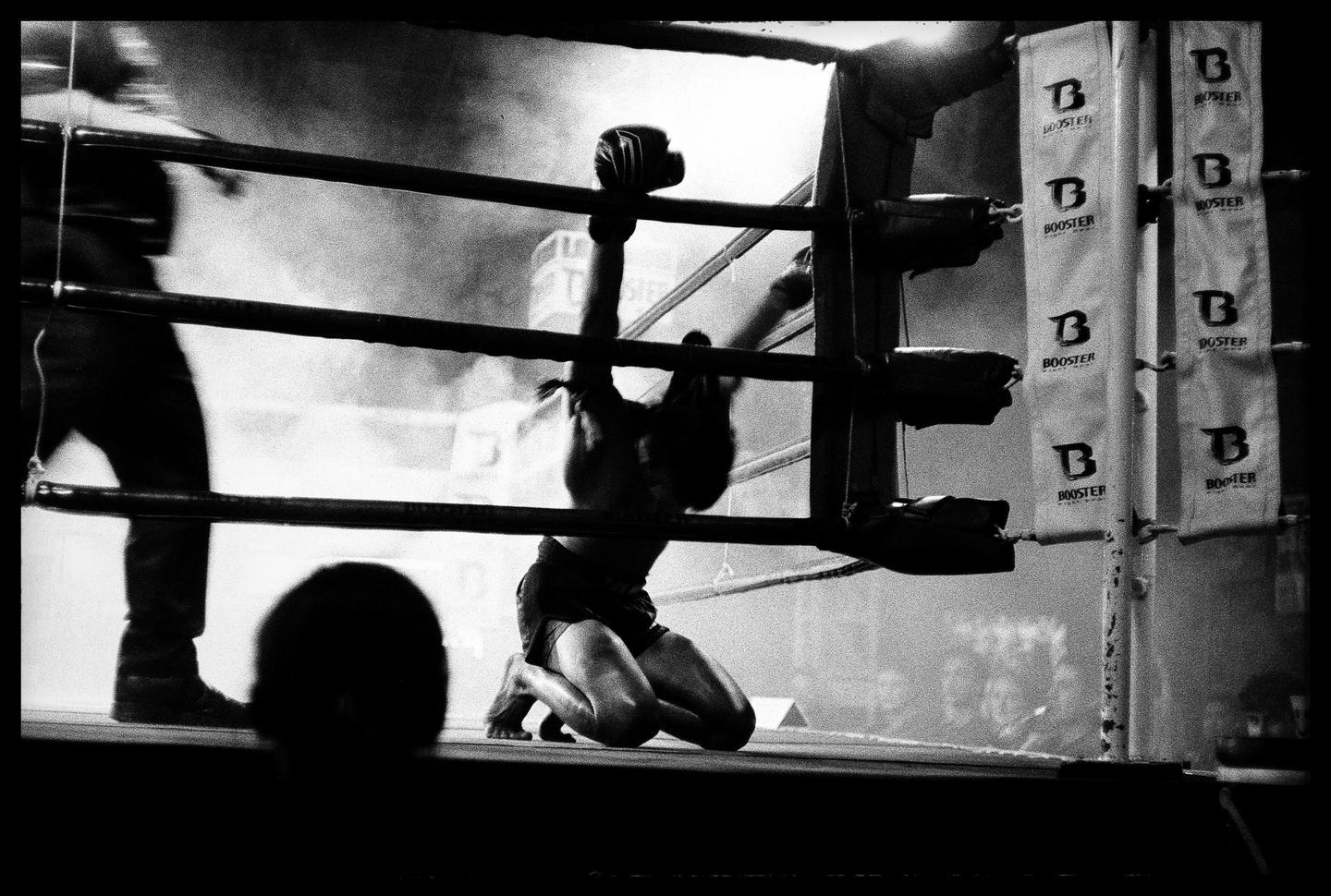 A muay thai fighter kneels in a boxing ring with their arms raised.