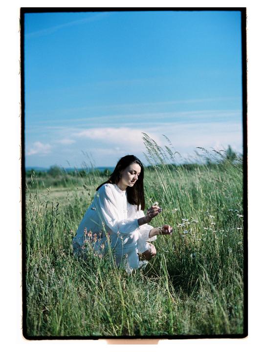 A woman in a white dress crouches in a field of tall grass and wildflowers under a clear blue sky.
