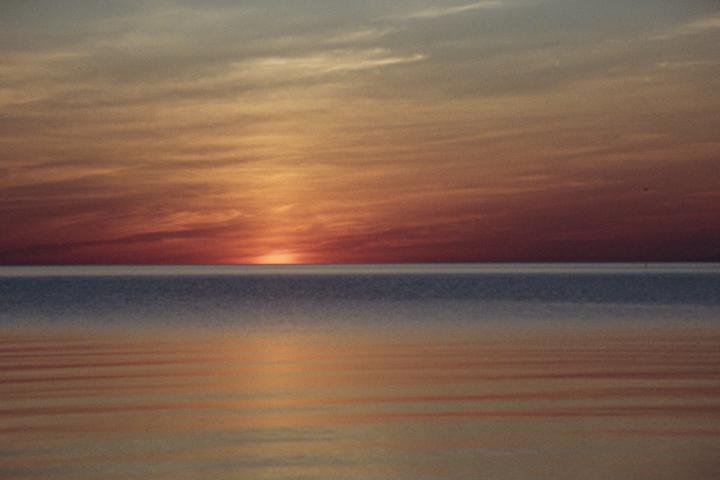 The sun sets over a body of water, casting an orange glow across the sky and reflecting on the water's surface.
