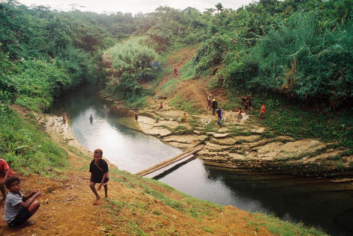 A group of people are gathered around a river in a lush, green landscape.