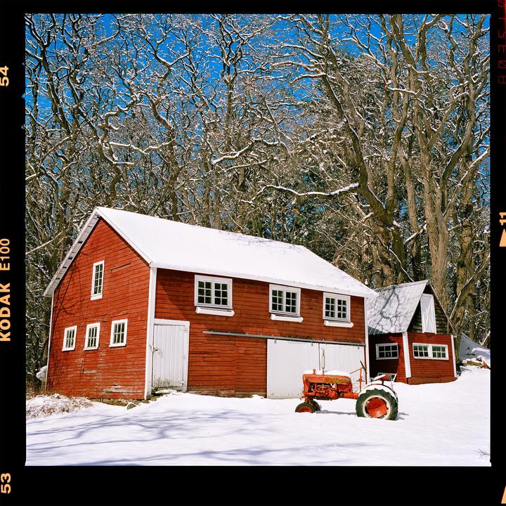 A red barn and a small red shed are covered in snow, with a red tractor parked in front of the barn, all set against a backdrop of snow-dusted trees under a clear blue sky.