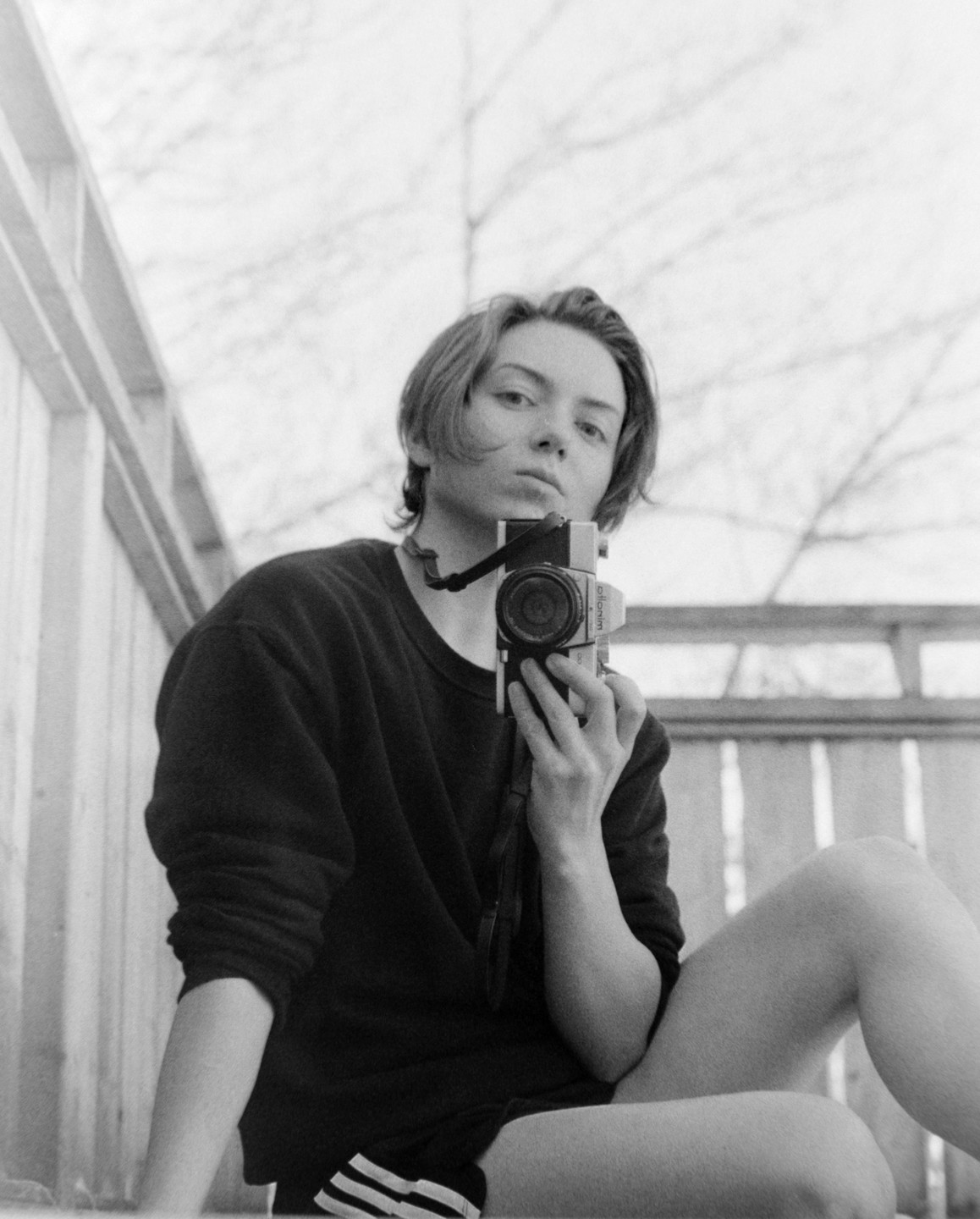 A young woman sits on a balcony holding a camera, looking directly at the viewer.