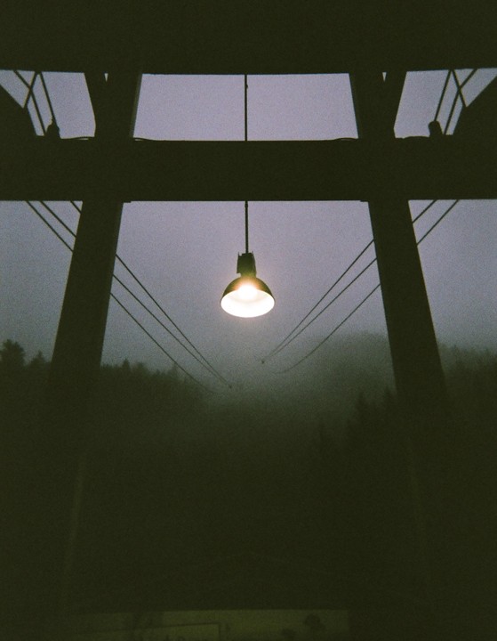 A single lightbulb hangs from a structure with cables extending into a foggy, tree-lined landscape.