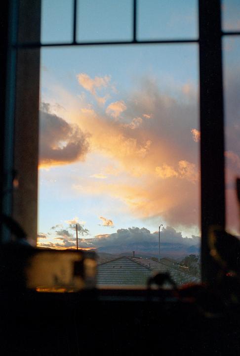 A window frames a dramatic sunset sky with orange and pink clouds over a distant mountain range and rooftops.