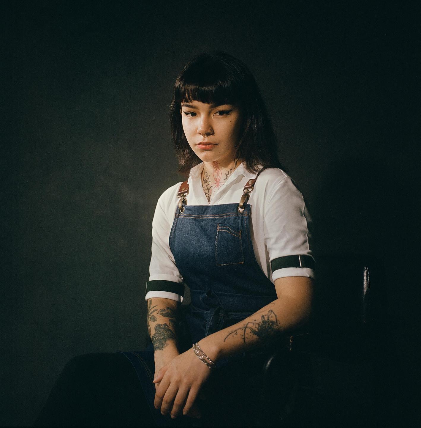 A woman with dark hair and tattoos sits wearing a denim apron over a white shirt.