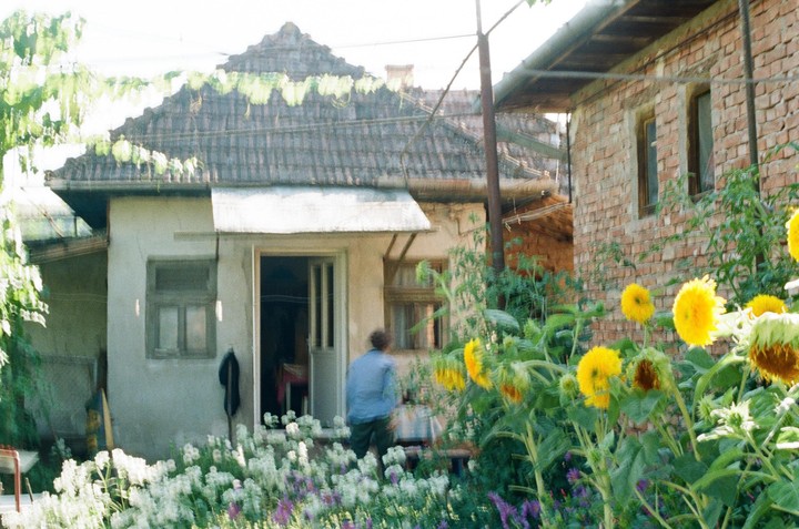 A man stands in the doorway of a house with a garden of sunflowers and white flowers in the foreground.