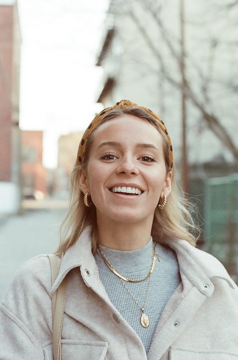 A young woman with a polka dot headband and gold jewelry smiles at the camera on a street.