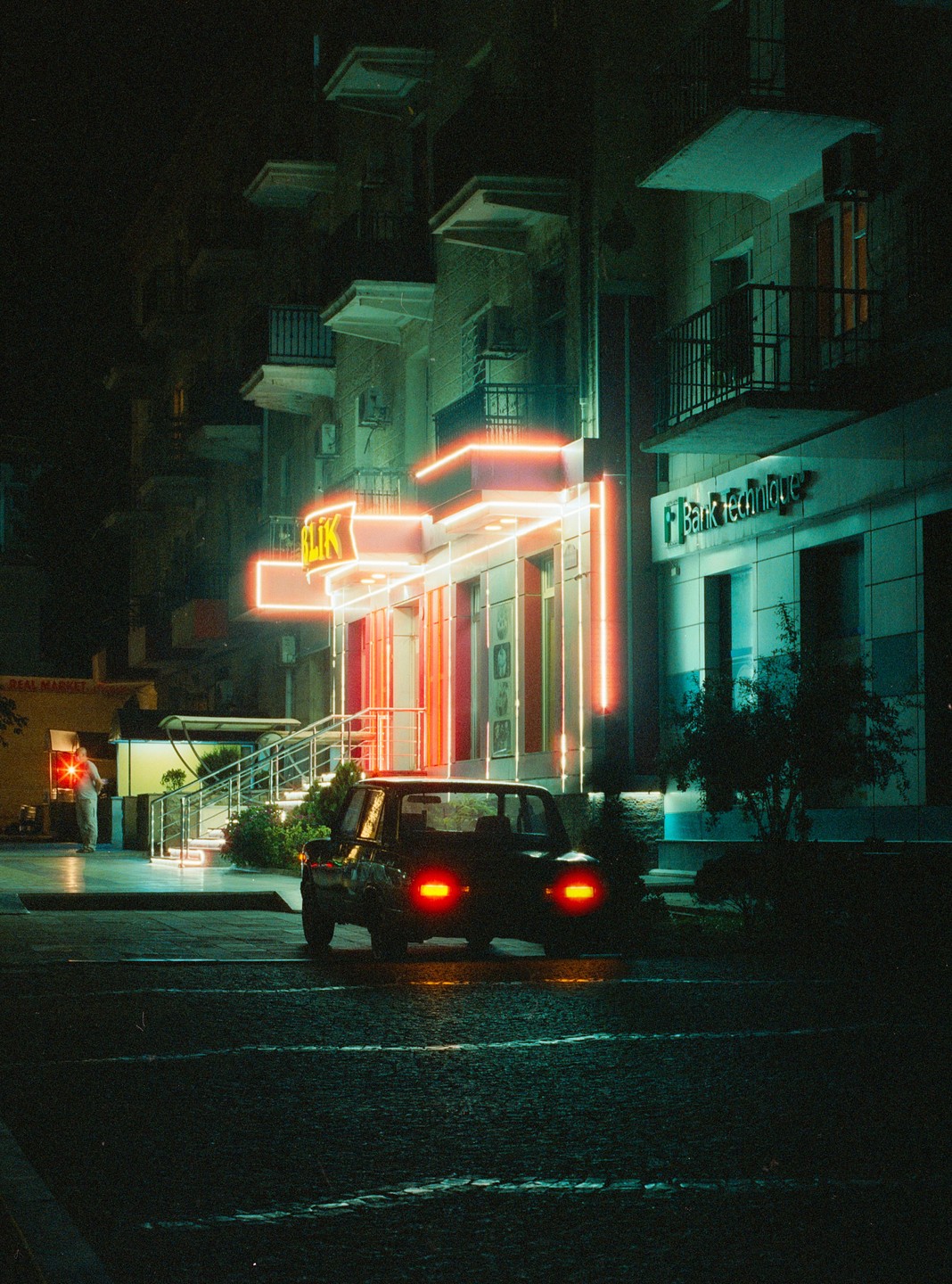 A car with illuminated taillights is parked on a street at night, with buildings featuring neon lights in the background.