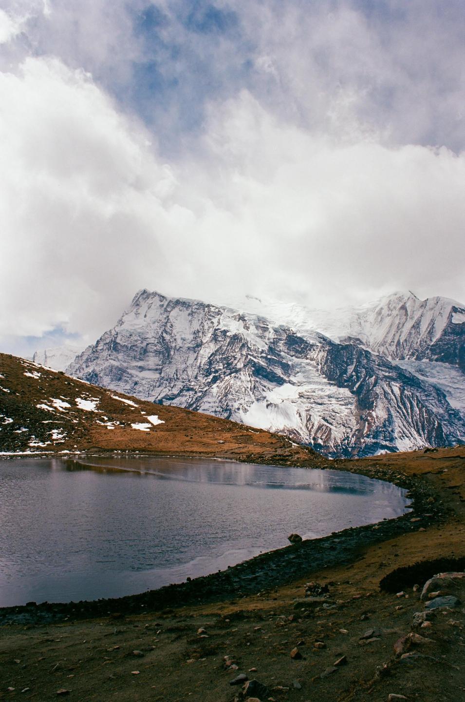 A large body of water sits in the foreground with snow-capped mountains and a cloudy sky in the background.
