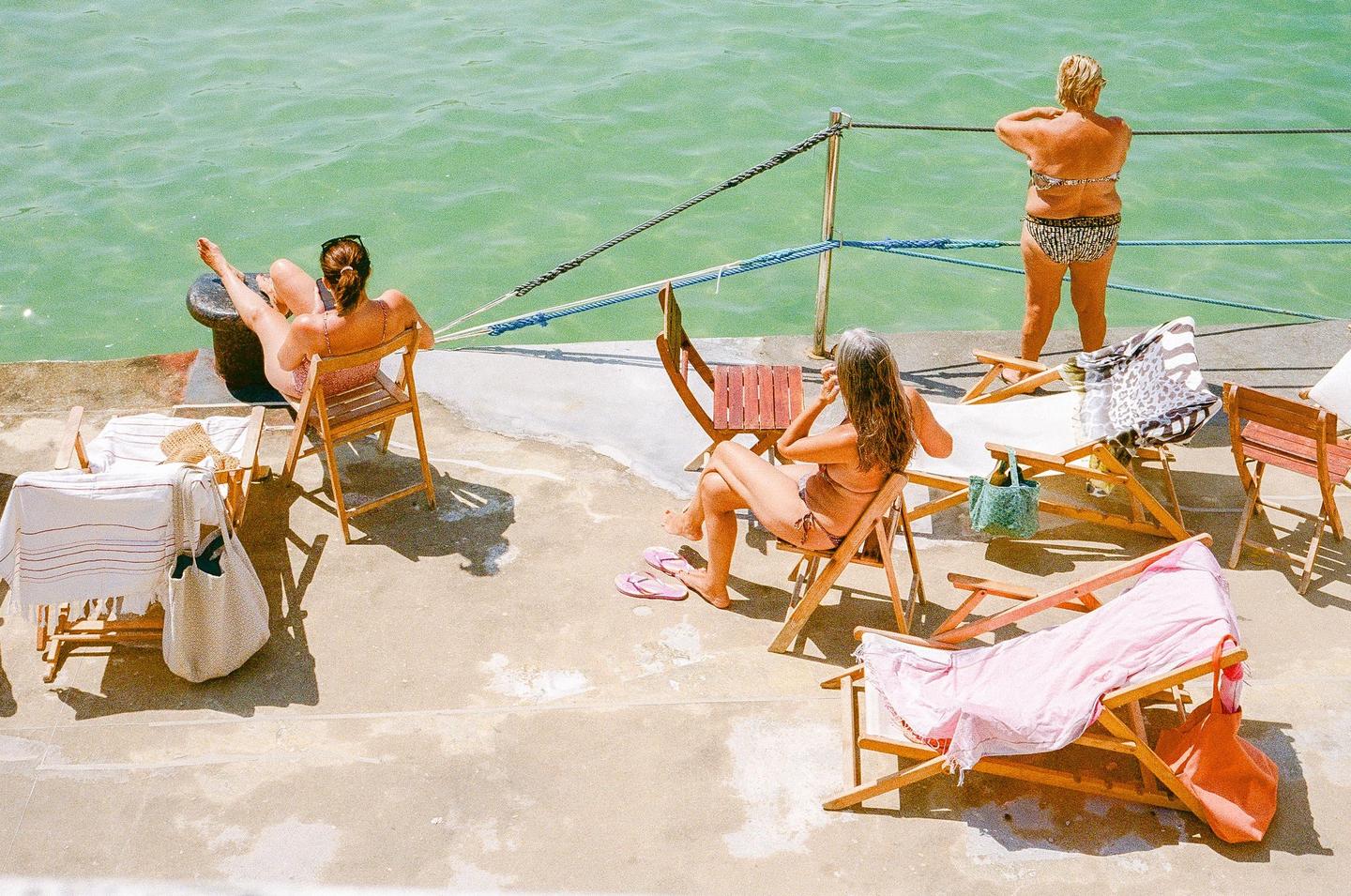 Three women are relaxing on a concrete pier next to green water on a sunny day.