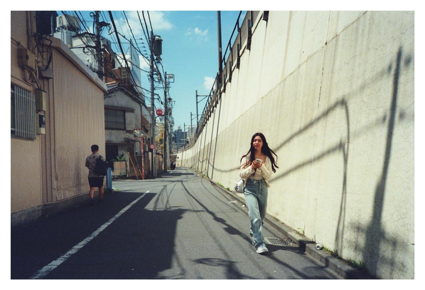 A woman walks down a narrow street in Japan, holding a phone, with a man walking ahead of her.