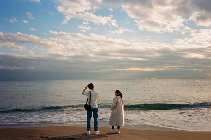 A couple stands on a sandy beach looking out at the ocean under a cloudy sky.