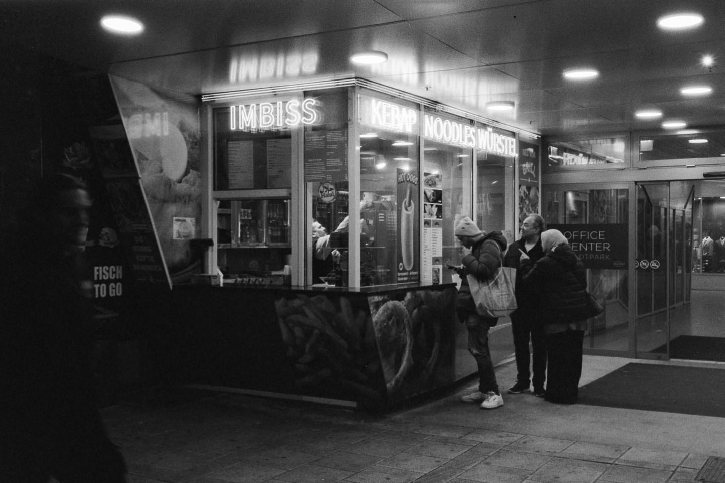 A group of people stand outside a brightly lit food stall at night.