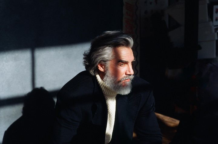 A man with gray hair and a beard looks to the right, illuminated by a shaft of sunlight.