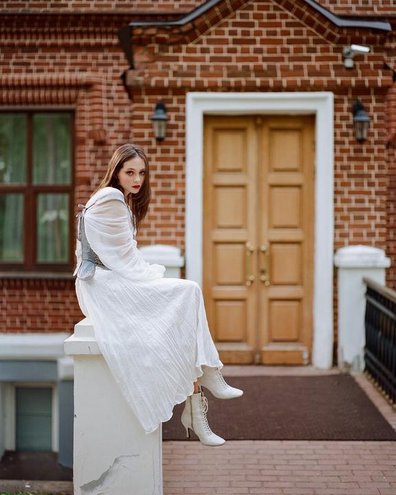 A woman in a white dress and boots sits on a white pillar in front of a brick building with a wooden door.