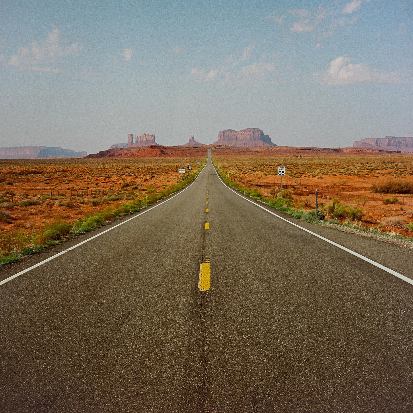 A long, straight road extends into the distance towards a series of mesas and buttes under a clear sky.