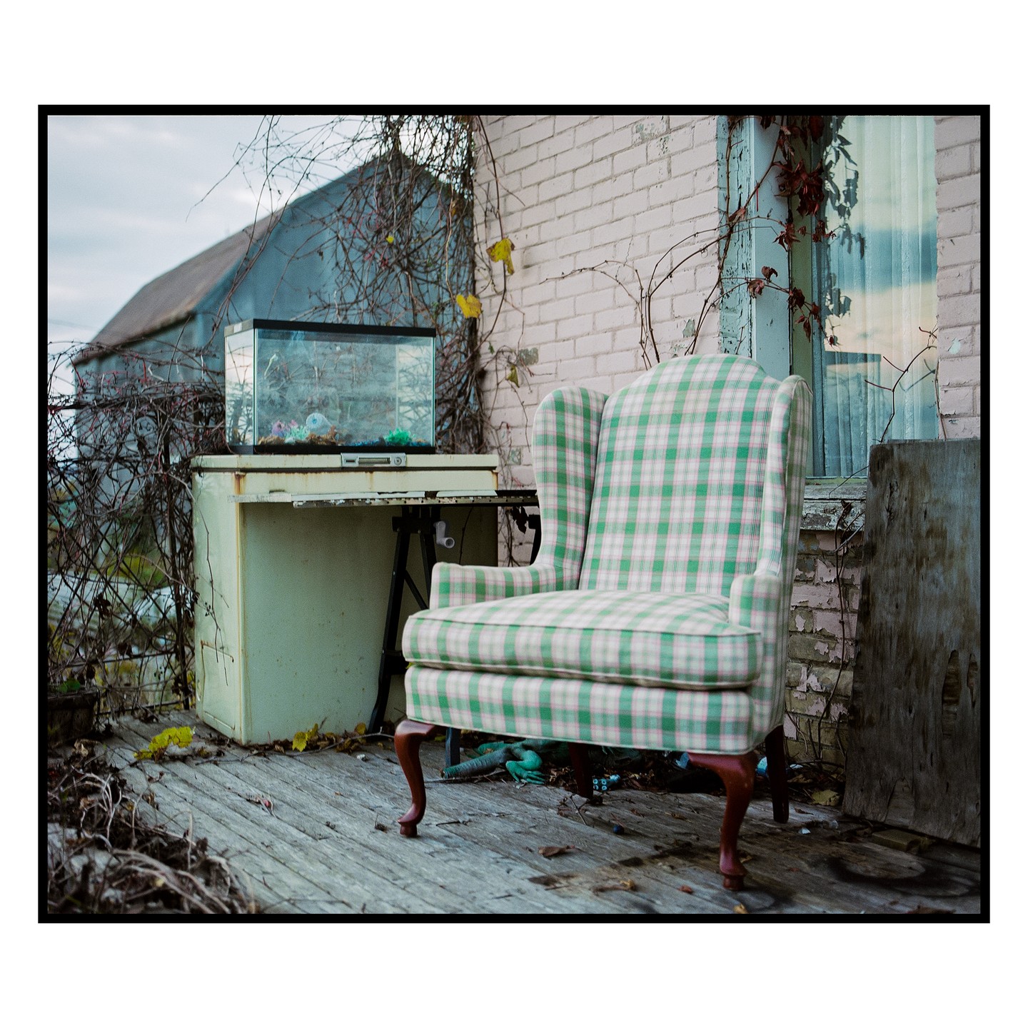 A green and white plaid armchair sits on a wooden porch next to a fish tank on a metal cabinet.