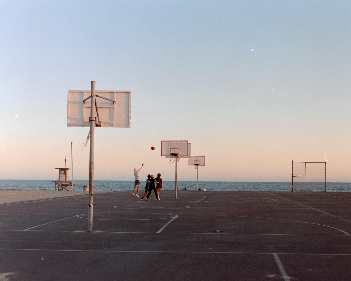 Three men play basketball on a court next to a beach with the ocean in the background at dusk.
