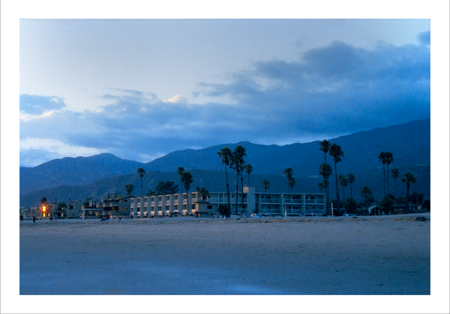 A beach with buildings and palm trees is visible in front of mountains under a cloudy sky at sunset.