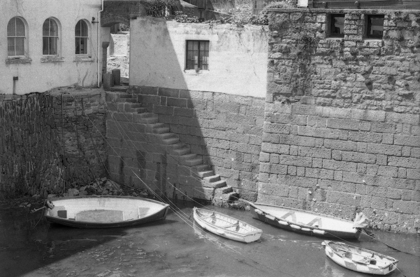 Four small boats are moored in the water next to a stone wall with steps leading up to a building.