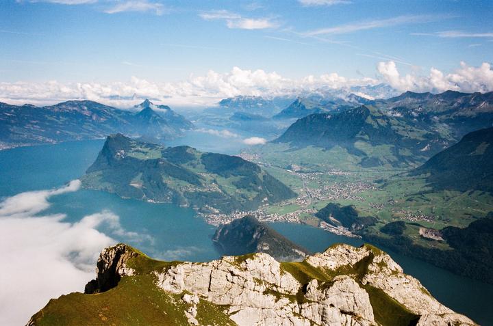 A high-angle landscape shows a lake surrounded by mountains and a small town, with clouds partially obscuring the view.