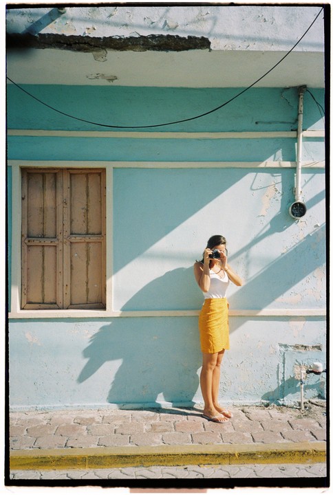 A woman in a white top and yellow skirt stands barefoot on a sidewalk, holding a camera to her face, in front of a light blue wall with a closed wooden window.