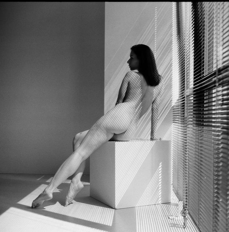 A nude woman sits on a white cube with light and shadow stripes cast across her body and the wall from a window blind.