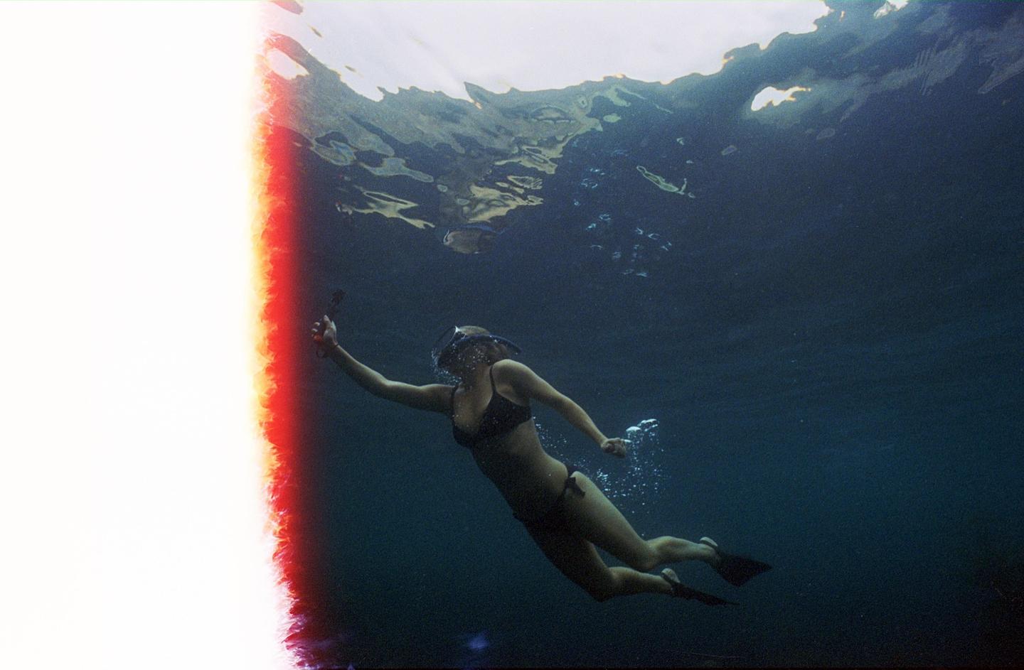 A woman in a bikini, snorkel, and fins swims underwater with a camera in her hand.