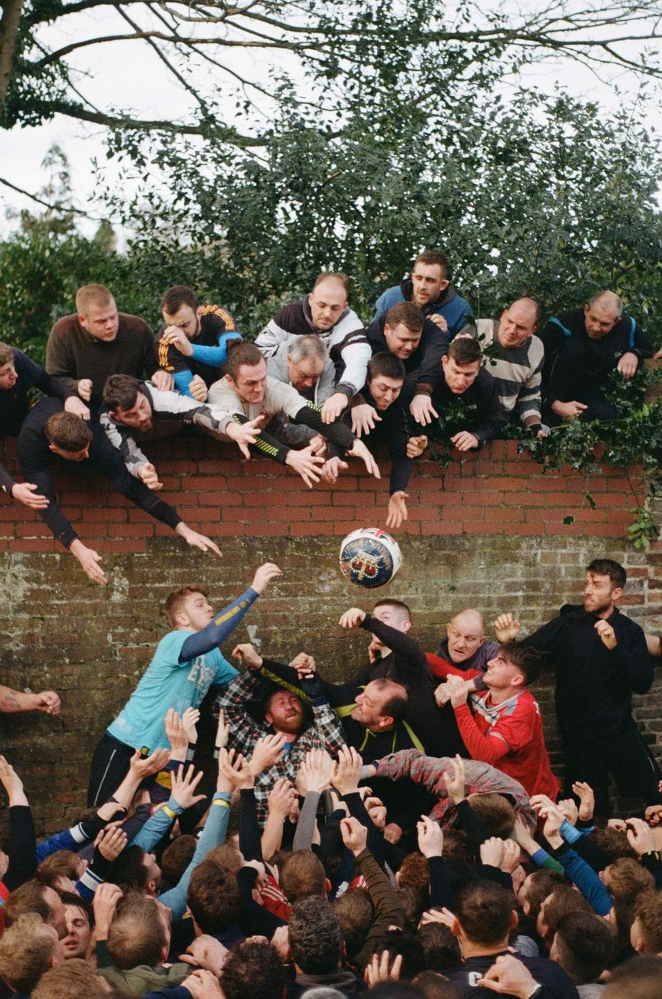 A large group of men are playing a game of Shrovetide football, with some men on a brick wall reaching for the ball and others in a scrum below.
