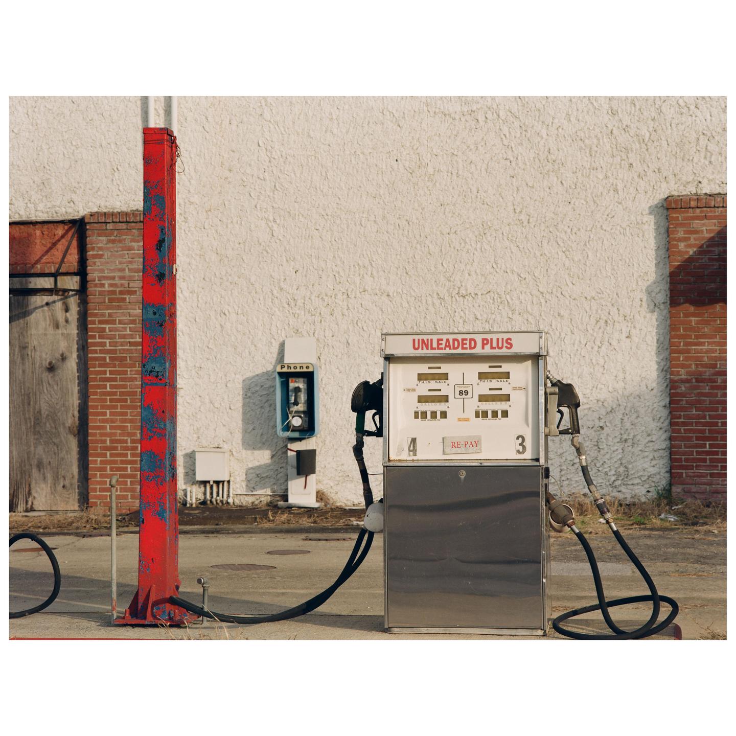 A gas pump and a payphone stand in front of a white building with a red pole in the foreground.