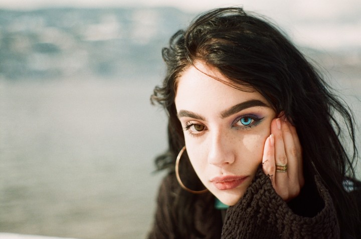 A woman with dark hair and heterochromia looks directly at the viewer, with one hand resting on her cheek.