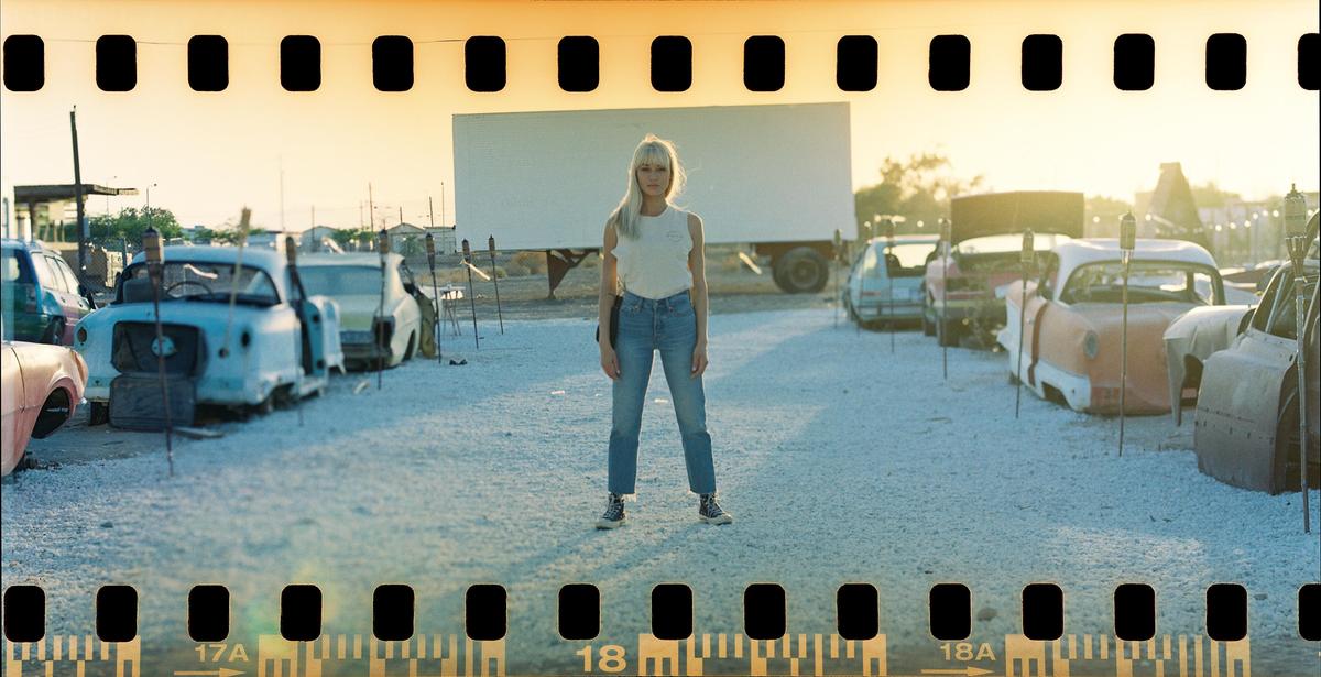 A woman stands in a gravel lot surrounded by old cars with a large white screen in the background.