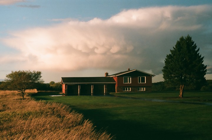 A red house with a long garage is situated between a field of tall grass and a manicured lawn, under a dramatic sky with large clouds.