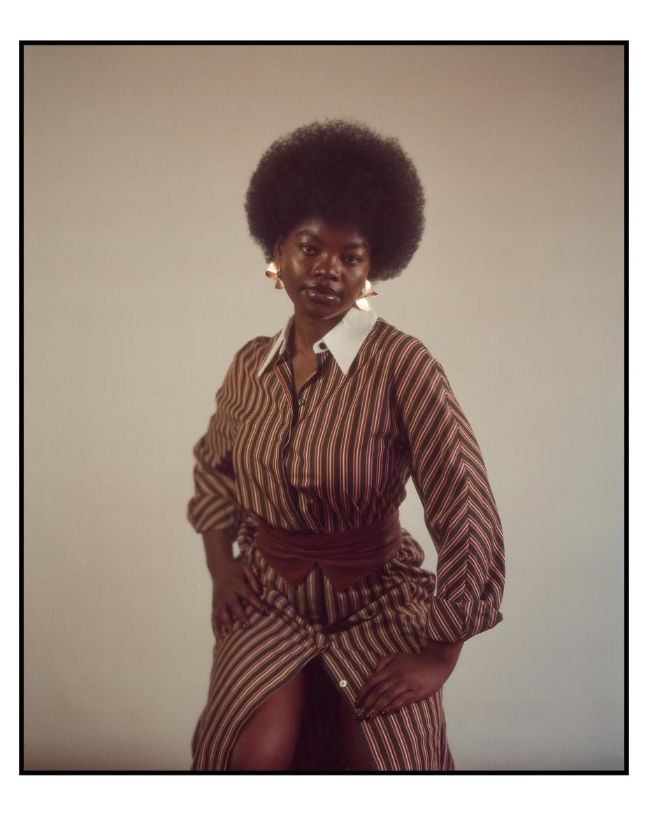 A woman with an afro wears a striped dress with a wide belt and large earrings.