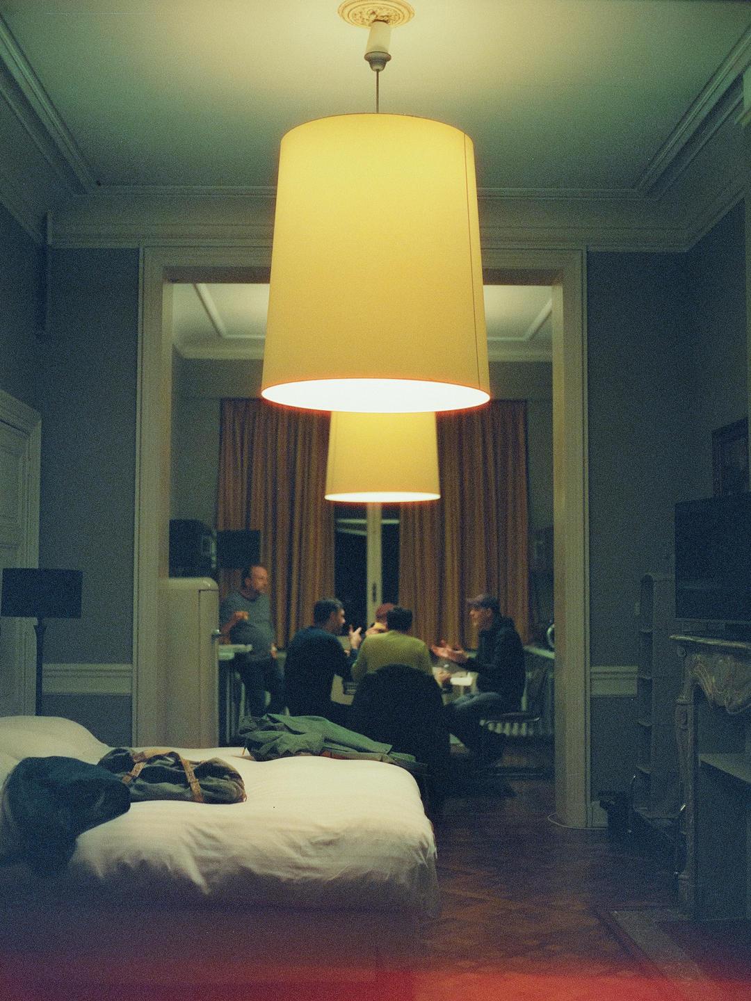 A bedroom with a bed in the foreground looks into another room where four men are gathered around a table.