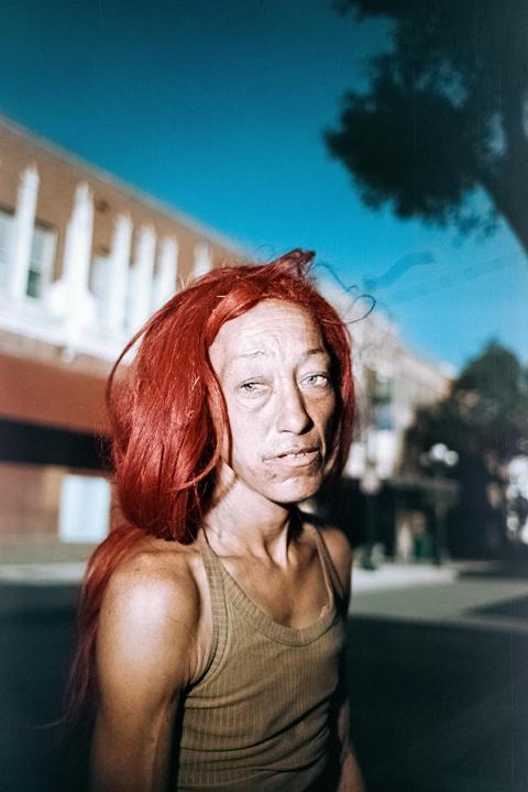 A woman with bright red hair and a tank top looks directly at the viewer on a street.