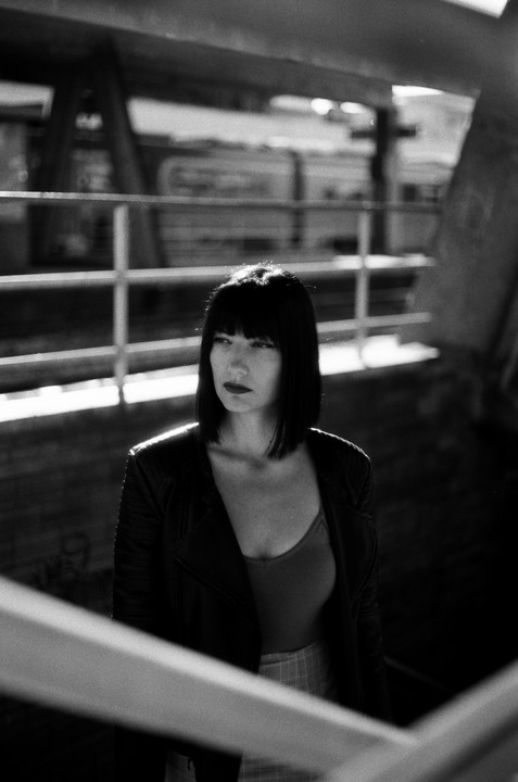 A woman with short dark hair and a leather jacket stands in a dark, industrial setting with railings.