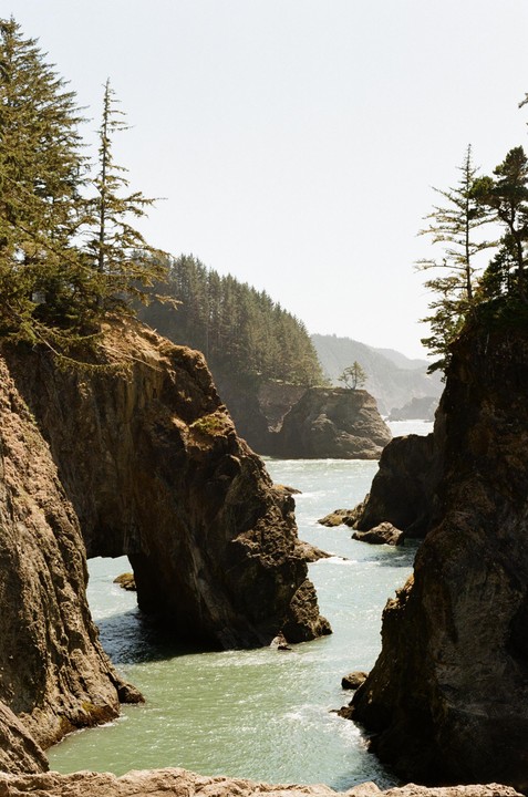 A narrow channel of water flows between two large rock formations with pine trees on top, leading to a forested coastline in the distance.